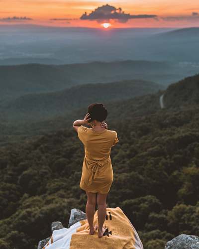 person woman in brown dress standing on cliff overlooking forest during golden hour outdoors