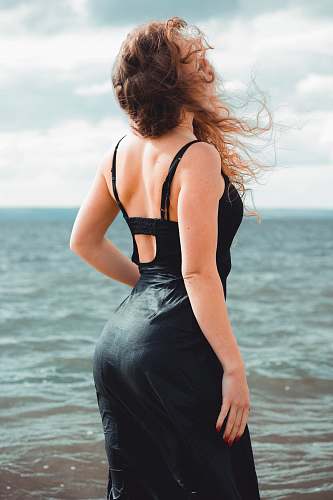 clothing woman wearing black spaghetti strap dress standing and facing on blue sea during daytime evening dress