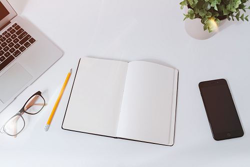 An open empty notebook on a white desk next to an iPhone and a MacBook