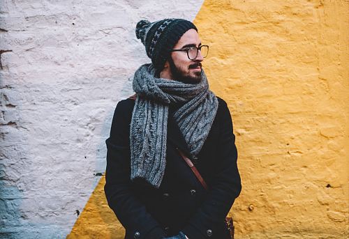 man in black coat with grey scarf standing on front of white and yellow painted wall