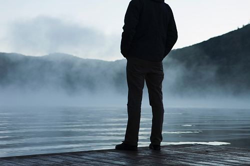 man standing facing body of water under white sky
