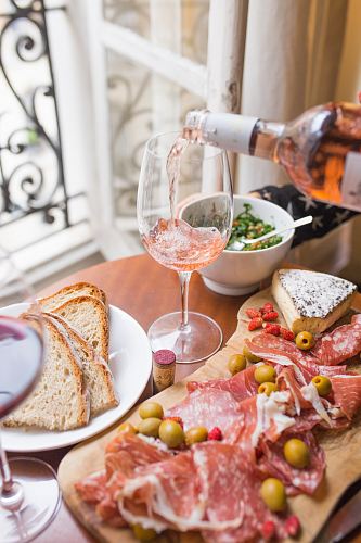 person pouring beverage on clear long-stem wine glass beside white ceramic plate with sliced bread and brown wooden board with bacons