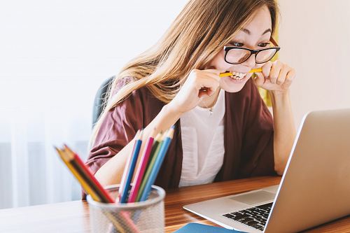 woman biting pencil while sitting on chair in front of computer during daytime