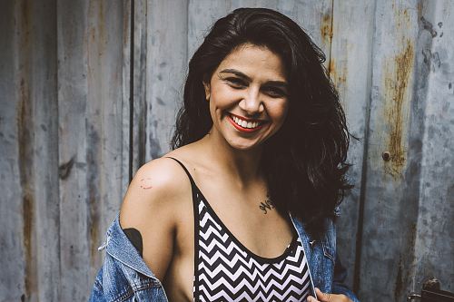 woman wearing white and black chevron print spaghetti-strap top with blue denim jacket standing behind gray wall photography