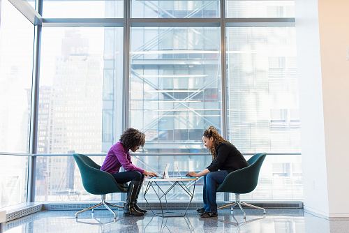 two women sits of padded chairs while using laptop computers