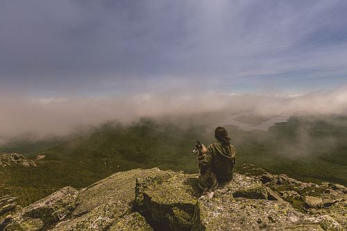 woman and dog sitting on gray concrete rock formation in front of mountain with fogs