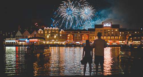  two people standing beside body of water watching fireworks 