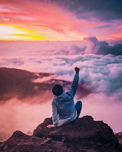 kailua-kona man sitting on mountain cliff facing white clouds rising one hand at golden hour united states
