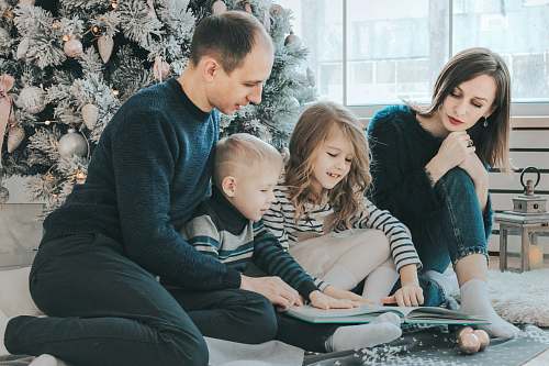 family girl and boy reading book sitting between man and woman beside Christmas tree person