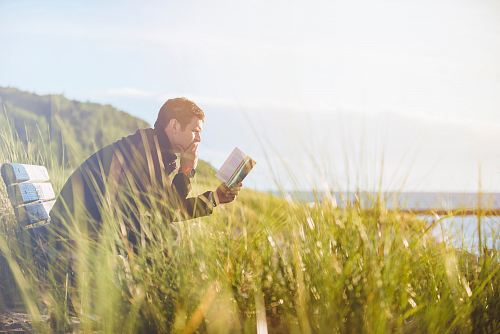 man reading book on beach near lake during daytime