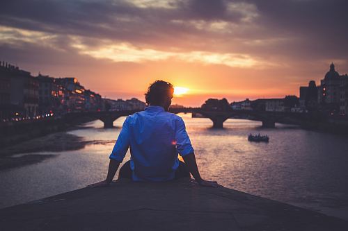 man wearing dress shirt sitting on dock near body of water during golden hour