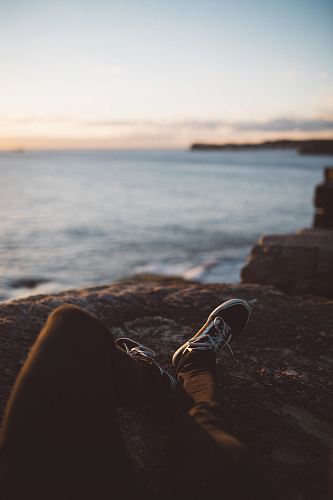 person wearing black pants and low-top sneakers lying down on brown rock formation near seashore