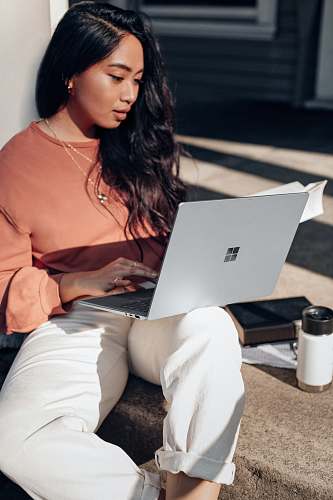 electronics woman in orange long sleeve shirt and white pants sitting on floor using Surface Laptop  pc